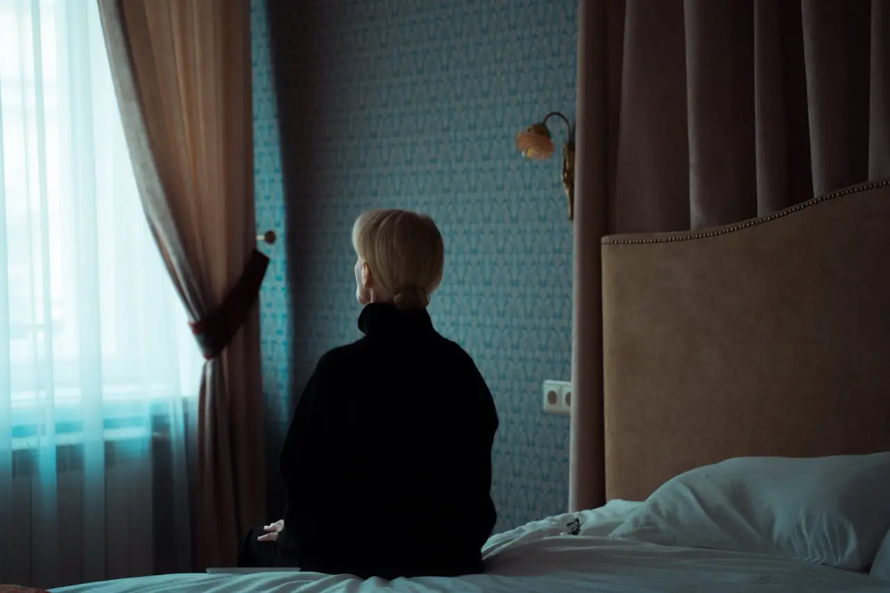 A woman in a dark room sits on a bed facing the window, reflecting solitude and contemplation.